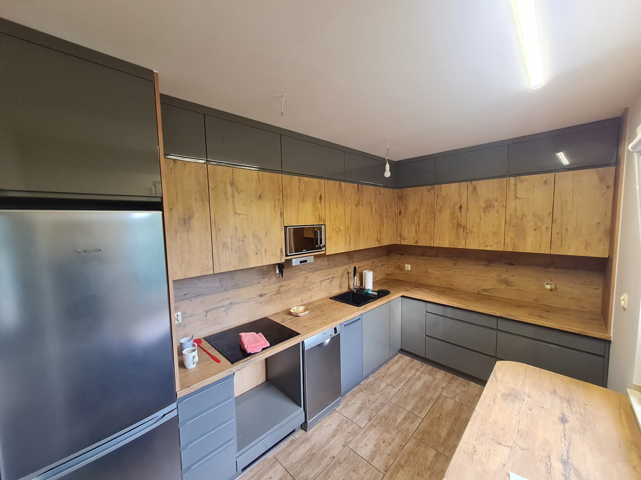 Nowoczesna kuchnia z szarymi szafkami dolnymi i drewnopodobnymi górnymi, srebrna lodówka Bosch po lewej, płyta indukcyjna z czerwoną ściereczką, jasna podłoga z płytek imitujących drewno.