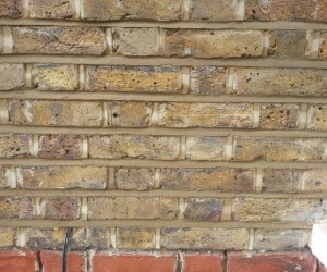 Fragment starego muru z cegły klinkierowej w odcieniach beżu i brązu, z widocznymi spoinami i drobnymi zabrudzeniami. W dolnej części kadru fragment czerwonej cegły.