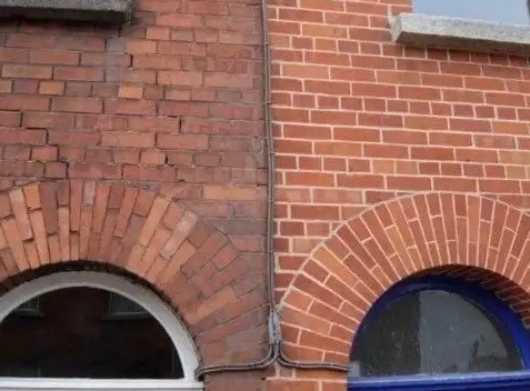 Fragment fasady budynku z czerwonej cegły, z widocznym efektem czyszczenia: lewa strona jest wyraźnie brudniejsza i ciemniejsza od prawej, która jest czysta i jaśniejsza. Widoczne okna z łukowymi...