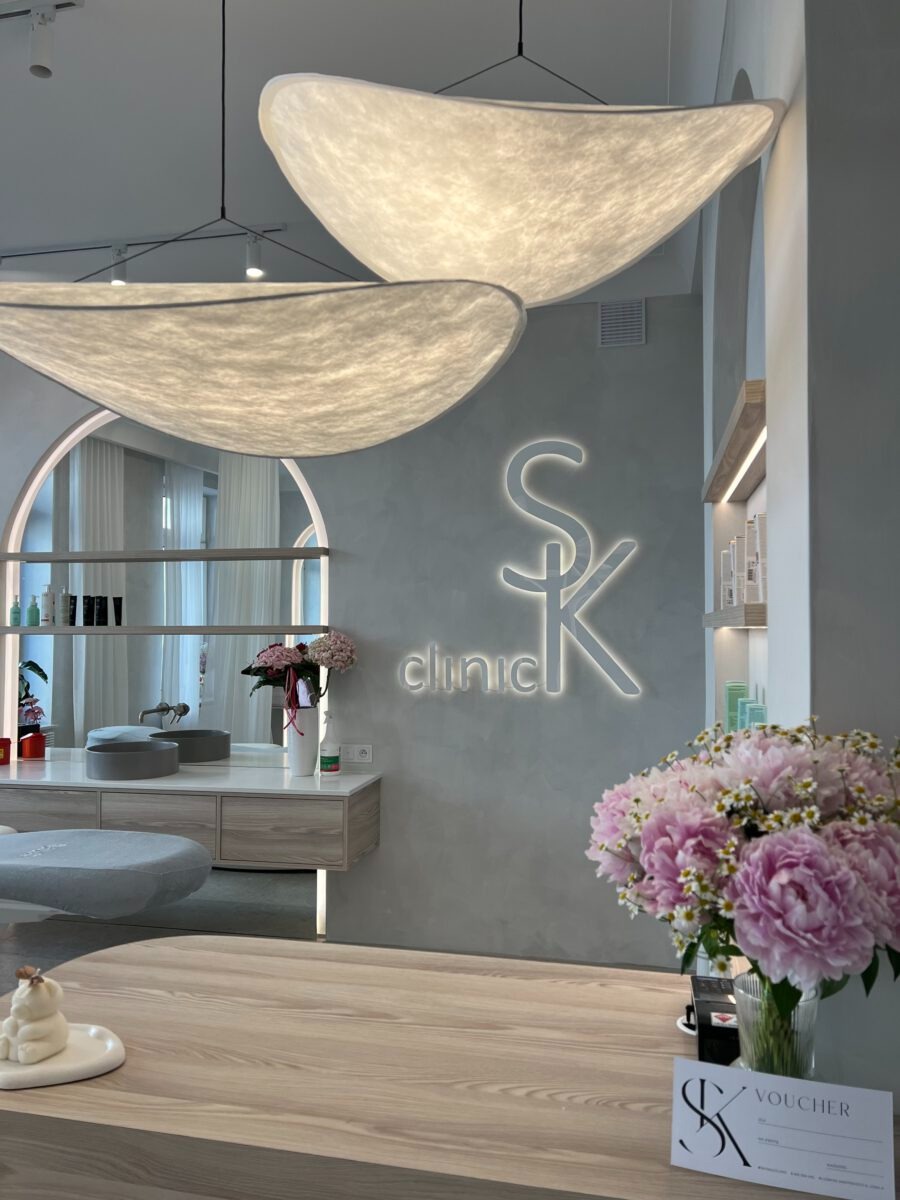 Jasne wnętrze kliniki z nowoczesnym oświetleniem w kształcie chmur, kwiatami w wazonie i logo firmy na ścianie, widok z recepcji.
