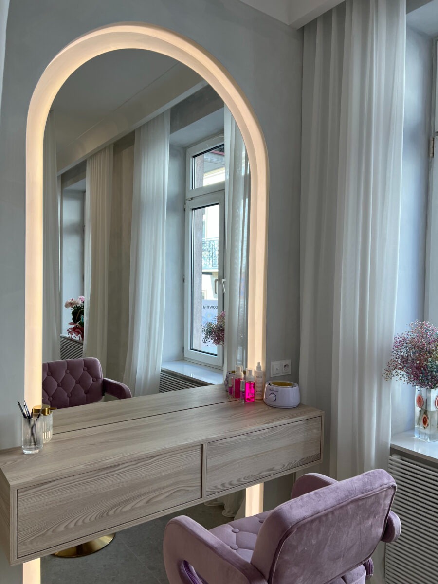 Jasne wnętrze salonu z toaletką i krzesłem w odcieniach fioletu, duże podświetlane lustro w kształcie łuku, kosmetyki na blacie, w tle okno z białymi zasłonami i bukiet kwiatów.