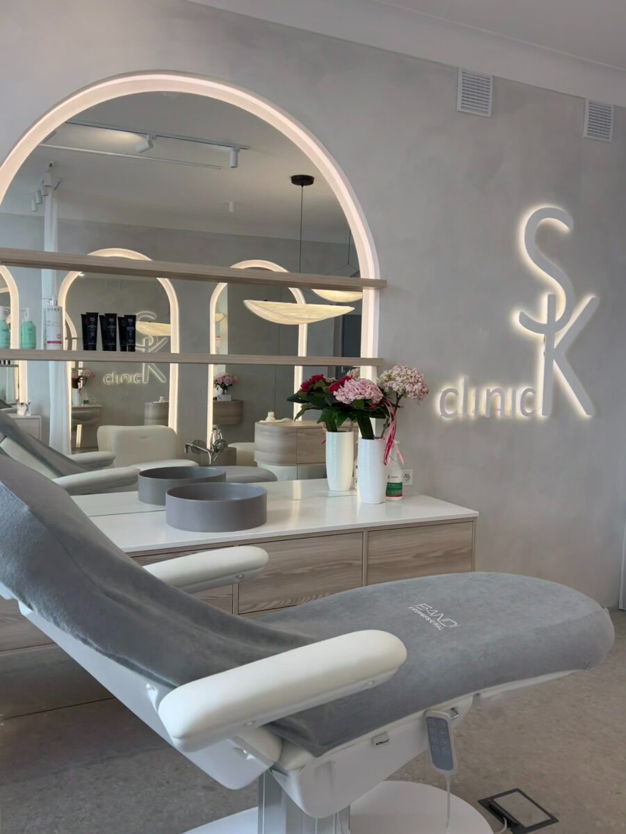 Jasne wnętrze gabinetu z fotelem zabiegowym przykrytym szarą narzutą, lustrem z podświetleniem łukowym i podświetlanym logo 'SK clinic' na ścianie. Na blacie wazon z kwiatami.