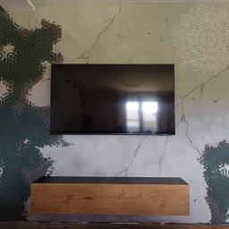 Patryk Radwański usługi budowlane - Ściana w salonie z dekoracyjnym tynkiem imitującym spękania, fragmenty zielonego wzoru geometrycznego i biała mandala, podwieszana szafka RTV z drewnianym frontem, wiszący telewizor.