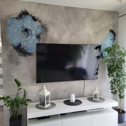 Patryk Radwański usługi budowlane - Szara ściana z efektem spękań i błękitnymi mandalami dekoracyjnymi za wiszącym telewizorem, biała szafka RTV z lampionami i roślinami doniczkowymi.
