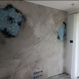 Patryk Radwański usługi budowlane - Ściana w salonie z dekoracyjnym tynkiem imitującym marmur, ozdobiona turkusowymi i szarymi wzorami mandali, z widocznymi gniazdkami elektrycznymi i fragmentem okna z białymi zasłonami.