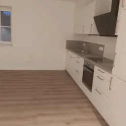 Nowoczesna, minimalistyczna kuchnia z białymi szafkami, szarym blatem i drewnianą podłogą. Widoczny czarny okap i okno z widokiem na ciemne niebo.