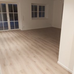 Patryk Radwański usługi budowlane - Jasny, pusty pokój z panelami podłogowymi w odcieniach beżu, białymi ścianami, oknami z białymi ramami i białymi drzwiami balkonowymi, widoczne gniazdka elektryczne przy podłodze.