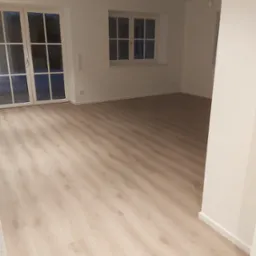 Jasny, pusty pokój z panelami podłogowymi w odcieniach beżu, białymi ścianami, oknami z białymi ramami i białymi drzwiami balkonowymi, widoczne gniazdka elektryczne przy podłodze.