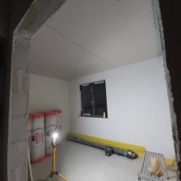 Patryk Radwański usługi budowlane - Pokój w trakcie prac wykończeniowych: ściany i sufit pokryte płytami kartonowo-gipsowymi, widoczne rolki wełny mineralnej Ursa, lampa robocza na trójnogu, drabina aluminiowa i poziomnica.
