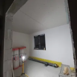 Pokój w trakcie prac wykończeniowych: ściany i sufit pokryte płytami kartonowo-gipsowymi, widoczne rolki wełny mineralnej Ursa, lampa robocza na trójnogu, drabina aluminiowa i poziomnica.