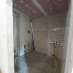 Wnętrze w trakcie remontu: położone płytki na ścianach i podłodze, sufit z płyt kartonowo-gipsowych z widocznymi łączeniami, prowizoryczne oświetlenie budowlane, wystające przewody i rury.