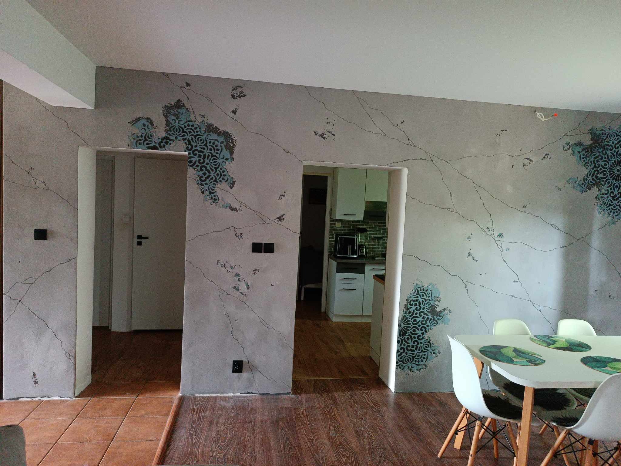 Wykończone wnętrze pokoju z dekoracyjną szarą ścianą imitującą spękania, ozdobioną niebiesko-czarnymi ornamentami, widoczne przejścia do innych pomieszczeń i stół z krzesłami.