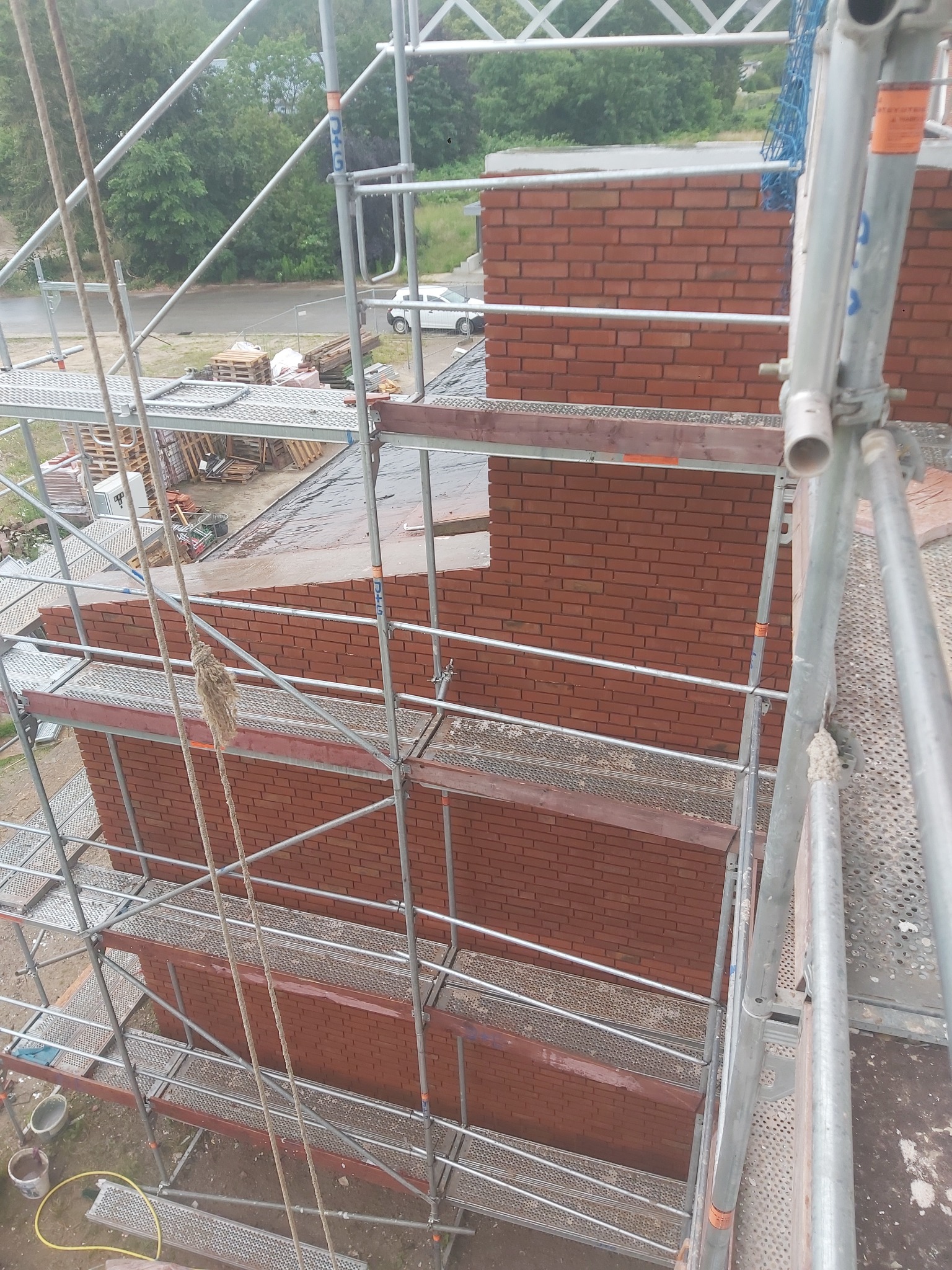 Wznoszenie muru z czerwonej cegły na zewnątrz budynku, widok z rusztowania, z widocznymi elementami konstrukcyjnymi i akcesoriami budowlanymi.