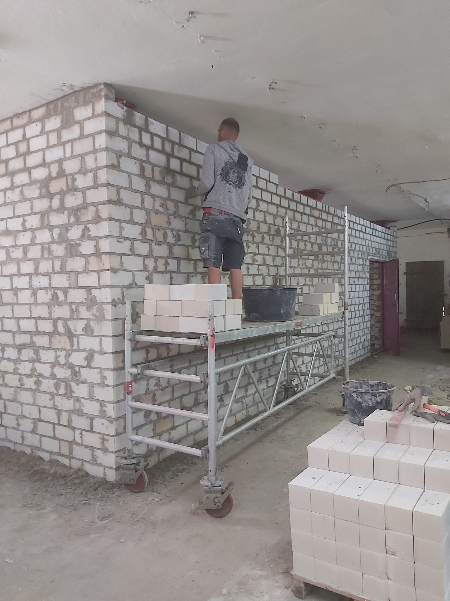 Murarz na rusztowaniu w trakcie wznoszenia ściany z białej cegły w surowym wnętrzu, widoczne zapasy cegieł i wiader z zaprawą.