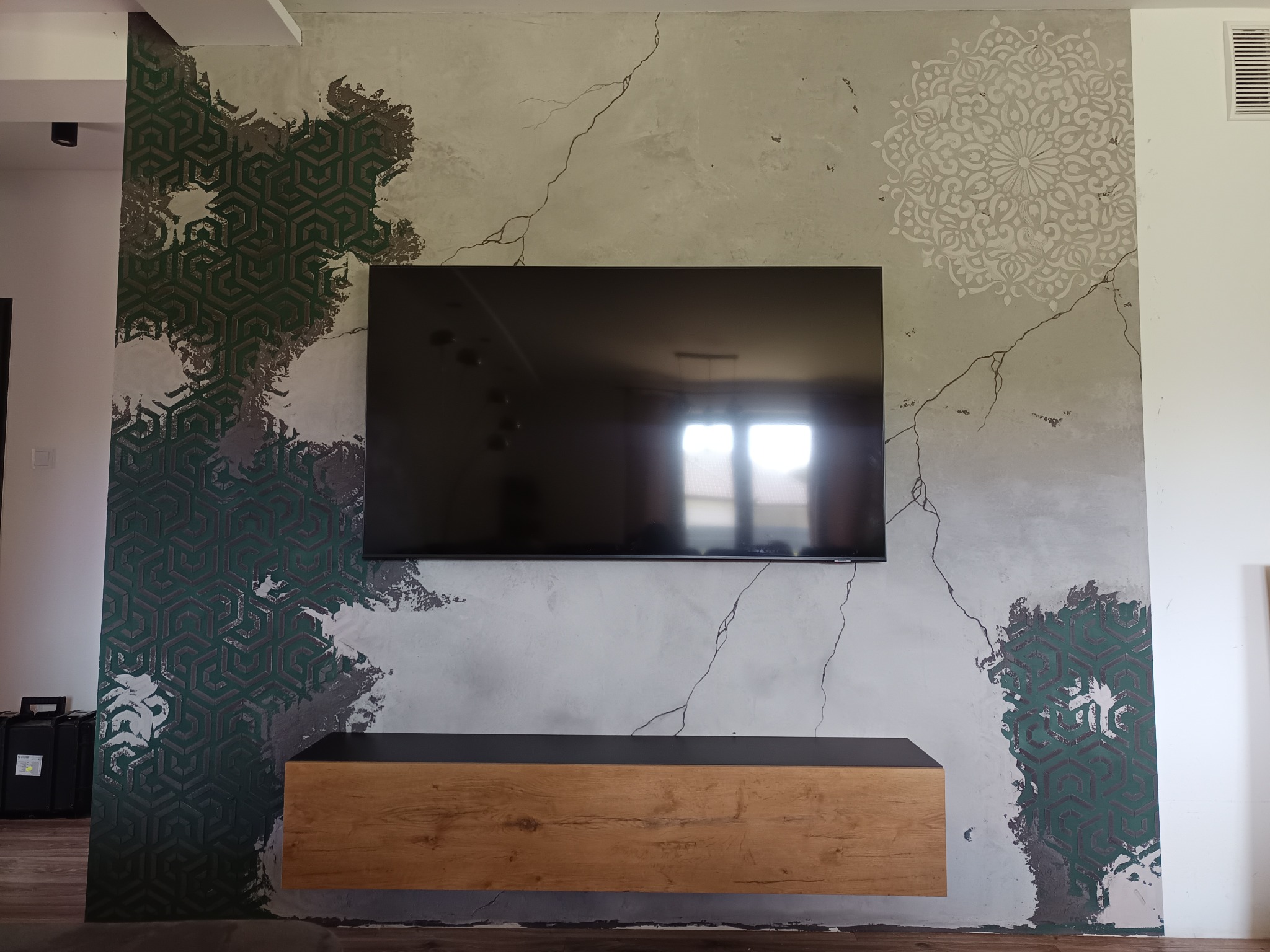 Ściana w salonie z dekoracyjnym tynkiem imitującym spękania, fragmenty zielonego wzoru geometrycznego i biała mandala, podwieszana szafka RTV z drewnianym frontem, wiszący telewizor.