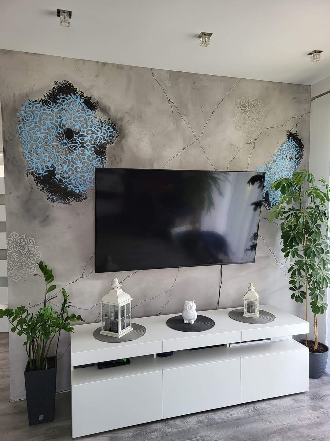 Szara ściana z efektem spękań i błękitnymi mandalami dekoracyjnymi za wiszącym telewizorem, biała szafka RTV z lampionami i roślinami doniczkowymi.