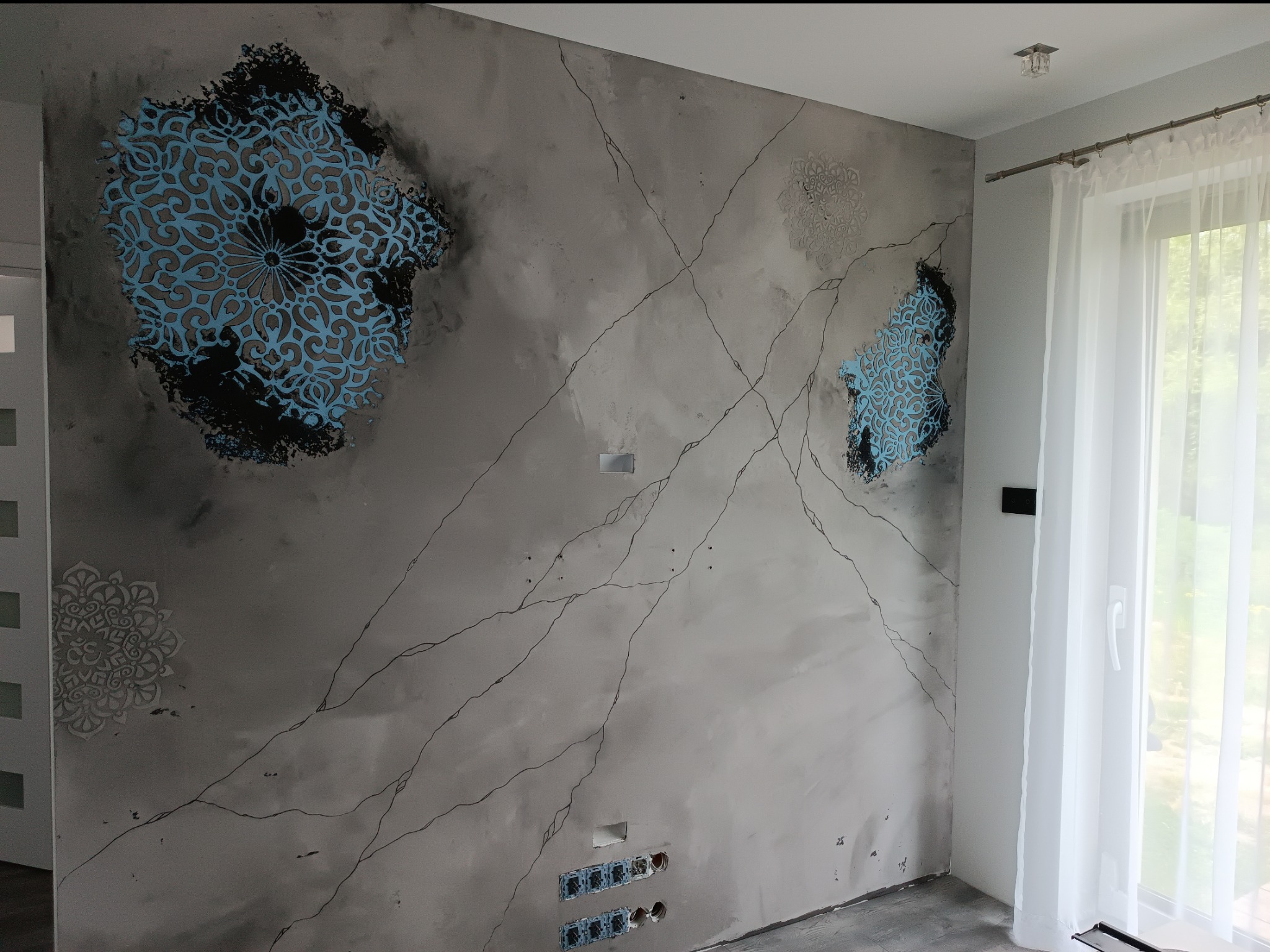 Ściana w salonie z dekoracyjnym tynkiem imitującym marmur, ozdobiona turkusowymi i szarymi wzorami mandali, z widocznymi gniazdkami elektrycznymi i fragmentem okna z białymi zasłonami.