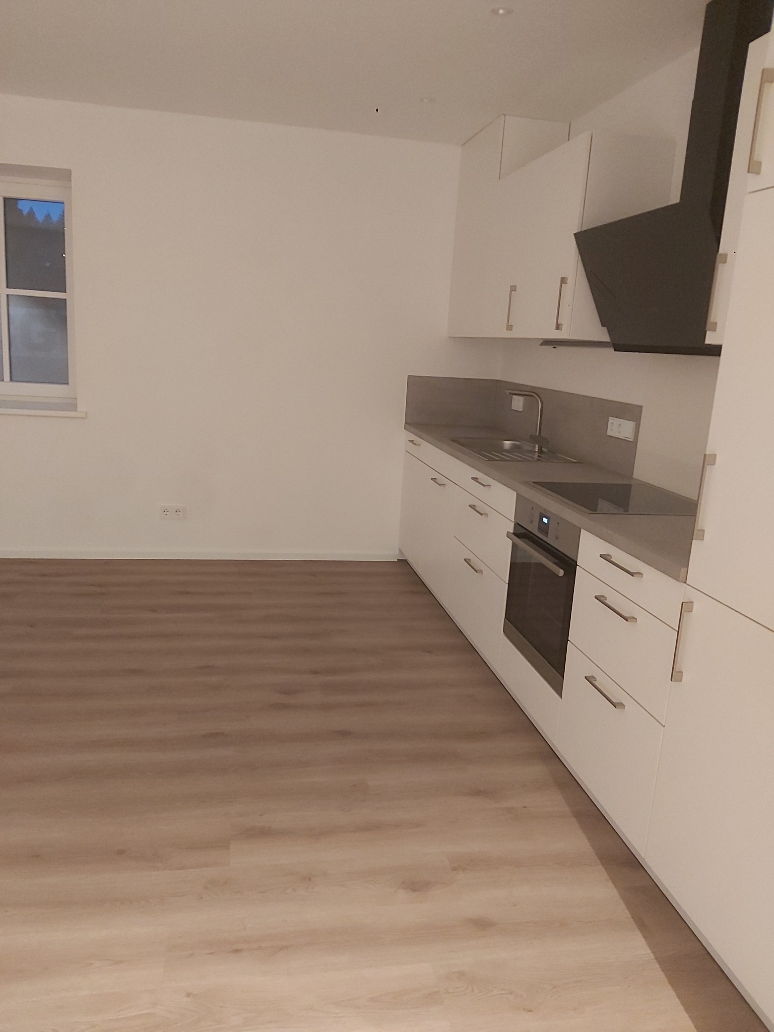 Nowoczesna, minimalistyczna kuchnia z białymi szafkami, szarym blatem i drewnianą podłogą. Widoczny czarny okap i okno z widokiem na ciemne niebo.