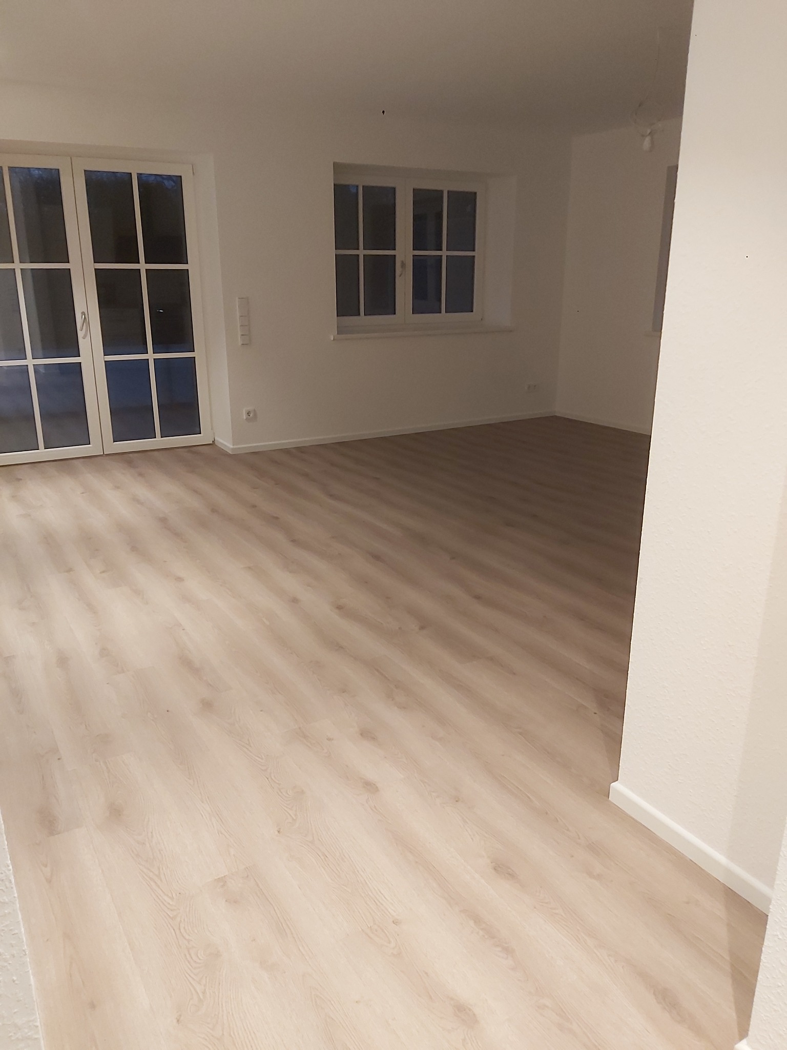 Jasny, pusty pokój z panelami podłogowymi w odcieniach beżu, białymi ścianami, oknami z białymi ramami i białymi drzwiami balkonowymi, widoczne gniazdka elektryczne przy podłodze.
