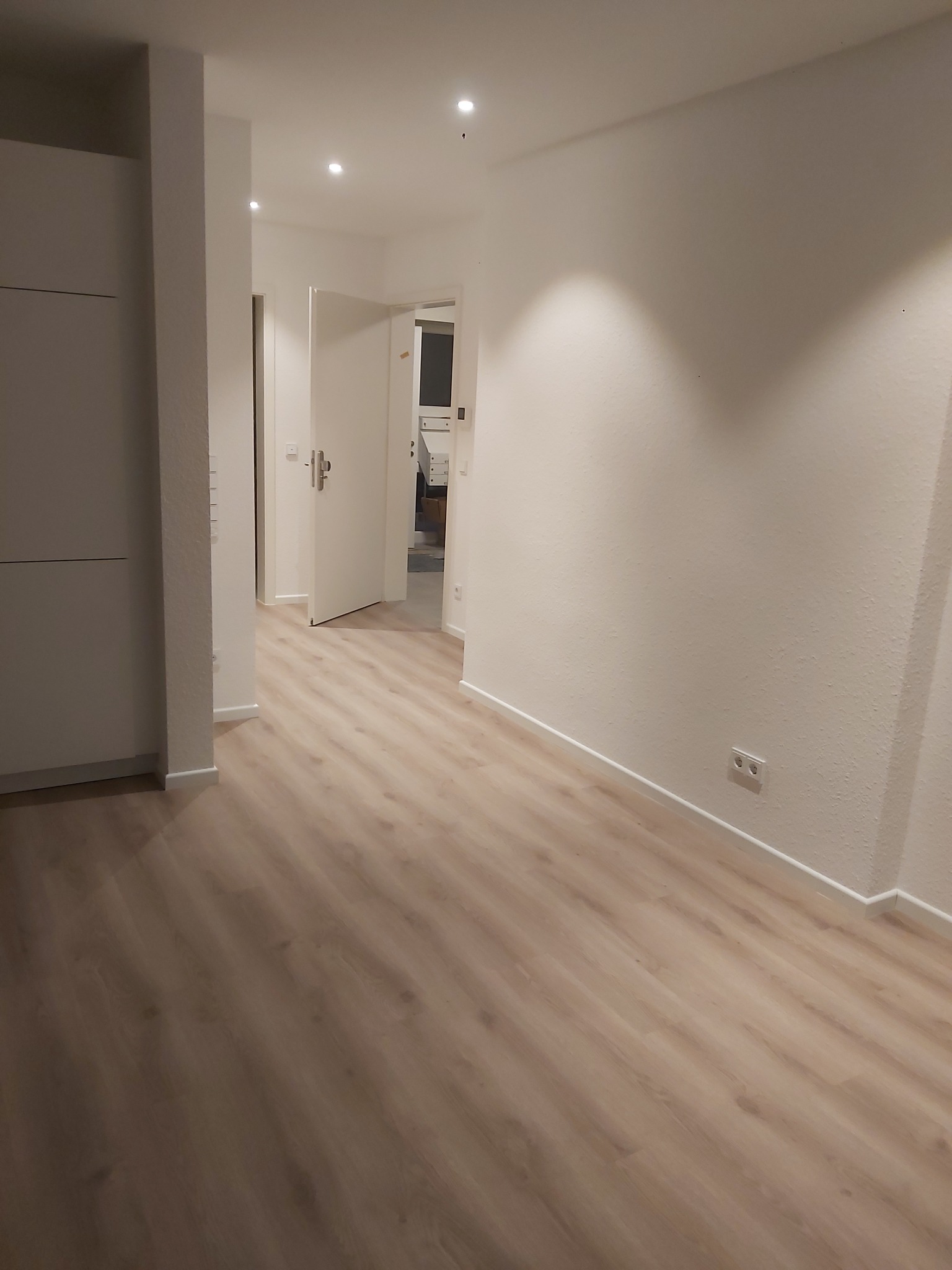 Nowo wykończone, puste wnętrze z panelami podłogowymi w odcieniach beżu, białe ściany z widocznymi gniazdkami elektrycznymi, otwarte drzwi prowadzące do kolejnego pomieszczenia, widoczna zabudowa...