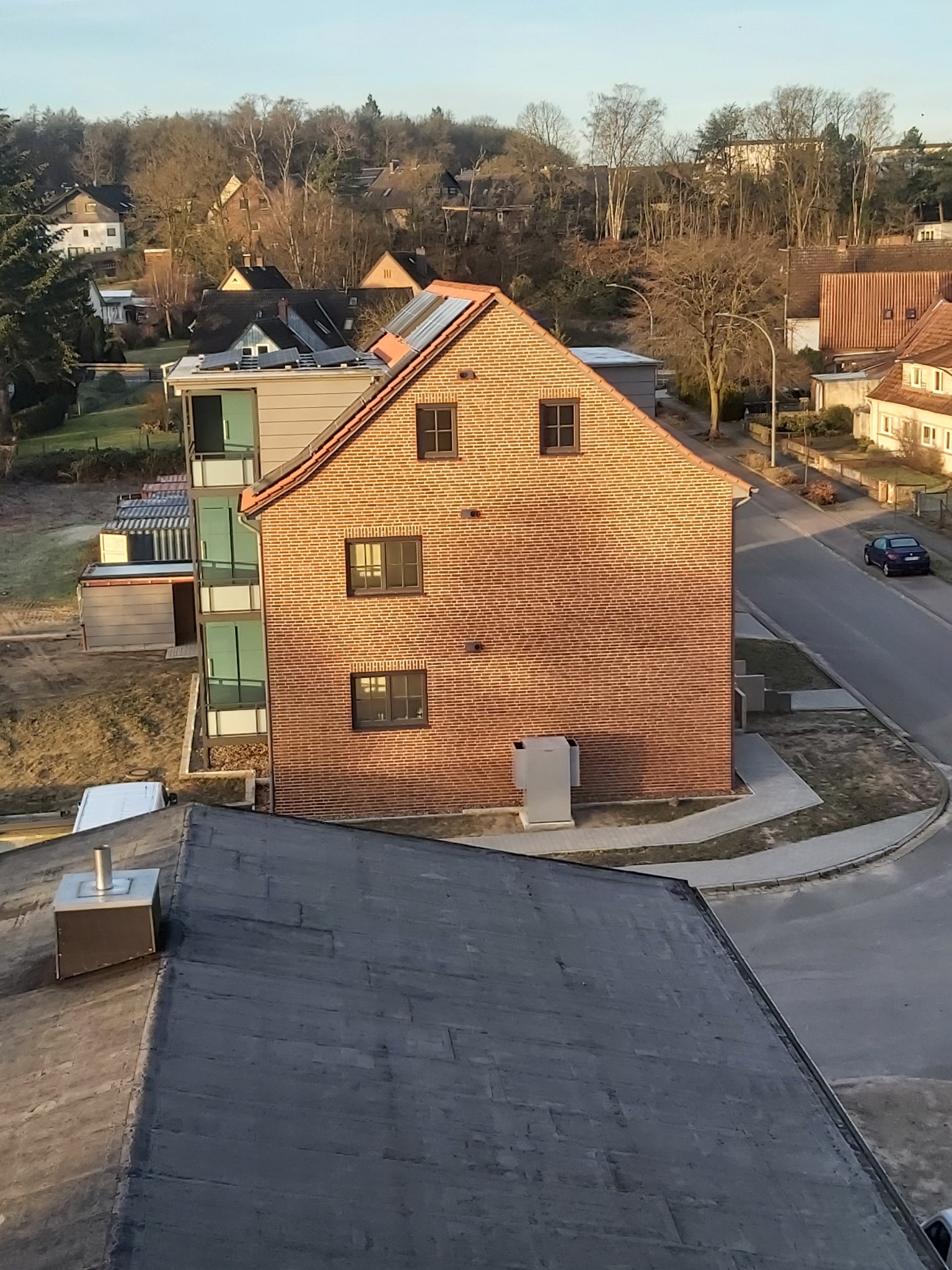 Widok z góry na ceglany budynek z oknami i balkonami, w tle zabudowa mieszkalna i drzewa, na pierwszym planie fragment dachu pokrytego papą.