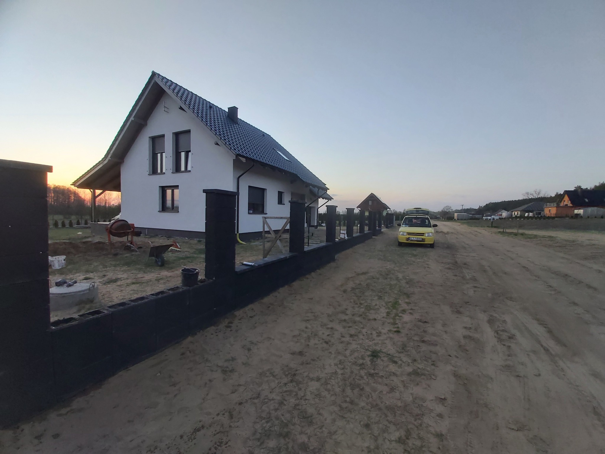 Nowy dom jednorodzinny z czarnym dachem i białymi ścianami, widoczny zza częściowo ukończonego czarnego ogrodzenia z betonowych bloczków, z betoniarką i żółtym samochodem w tle.