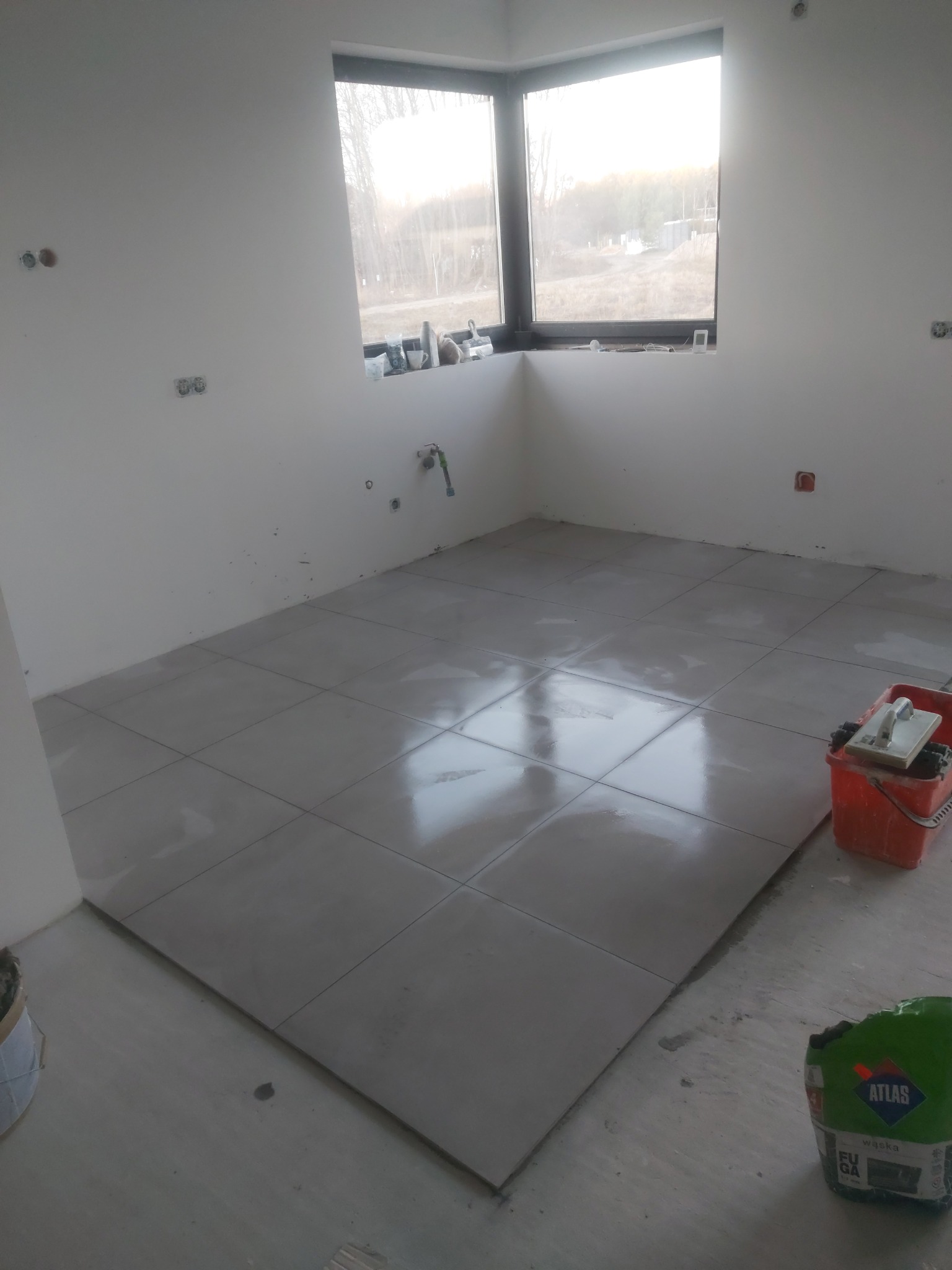 Układanie jasnoszarych płytek podłogowych w surowym wnętrzu z oknem, widoczny worek z fugą Atlas i wiadro z narzędziami do fugowania.