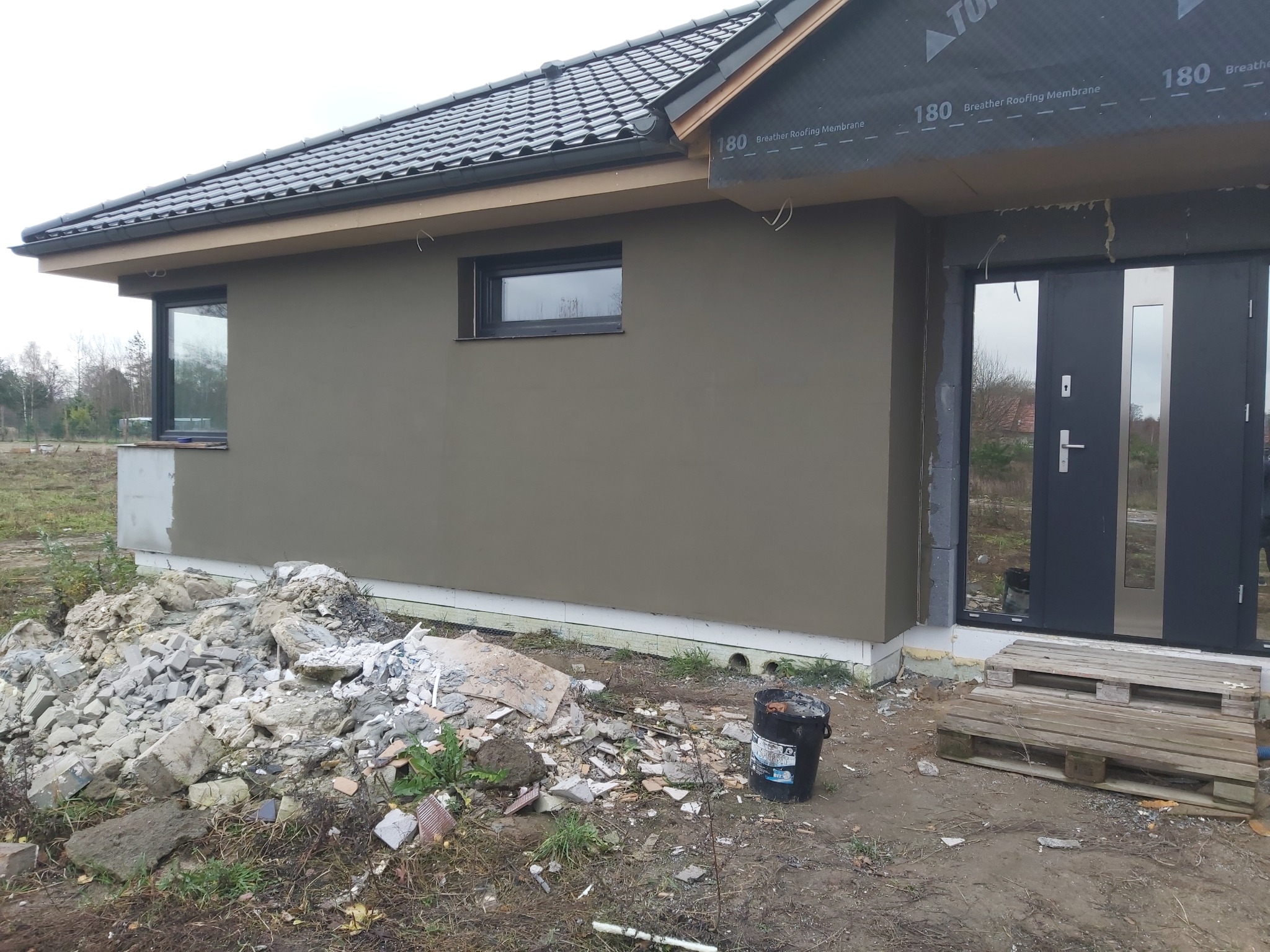 Dom jednorodzinny w trakcie prac wykończeniowych elewacji, widoczny stos gruzu i wiadro po farbie, okna z czarnymi ramami, częściowo zabezpieczone wejście, dach pokryty dachówką.