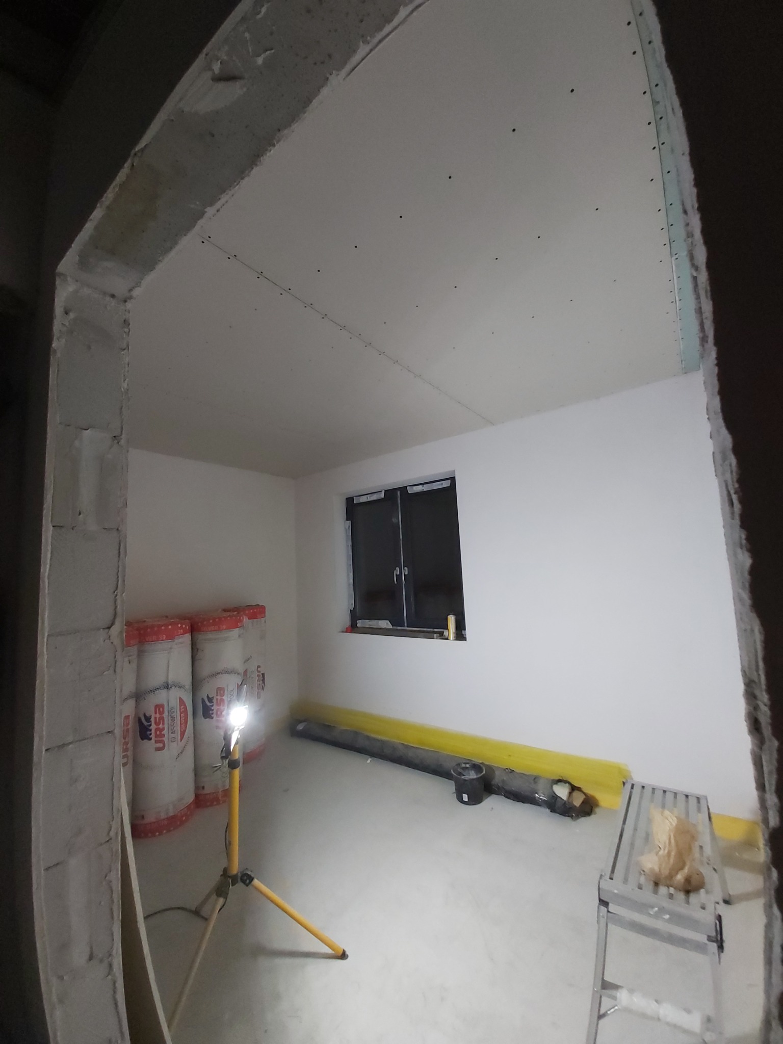 Pokój w trakcie prac wykończeniowych: ściany i sufit pokryte płytami kartonowo-gipsowymi, widoczne rolki wełny mineralnej Ursa, lampa robocza na trójnogu, drabina aluminiowa i poziomnica.
