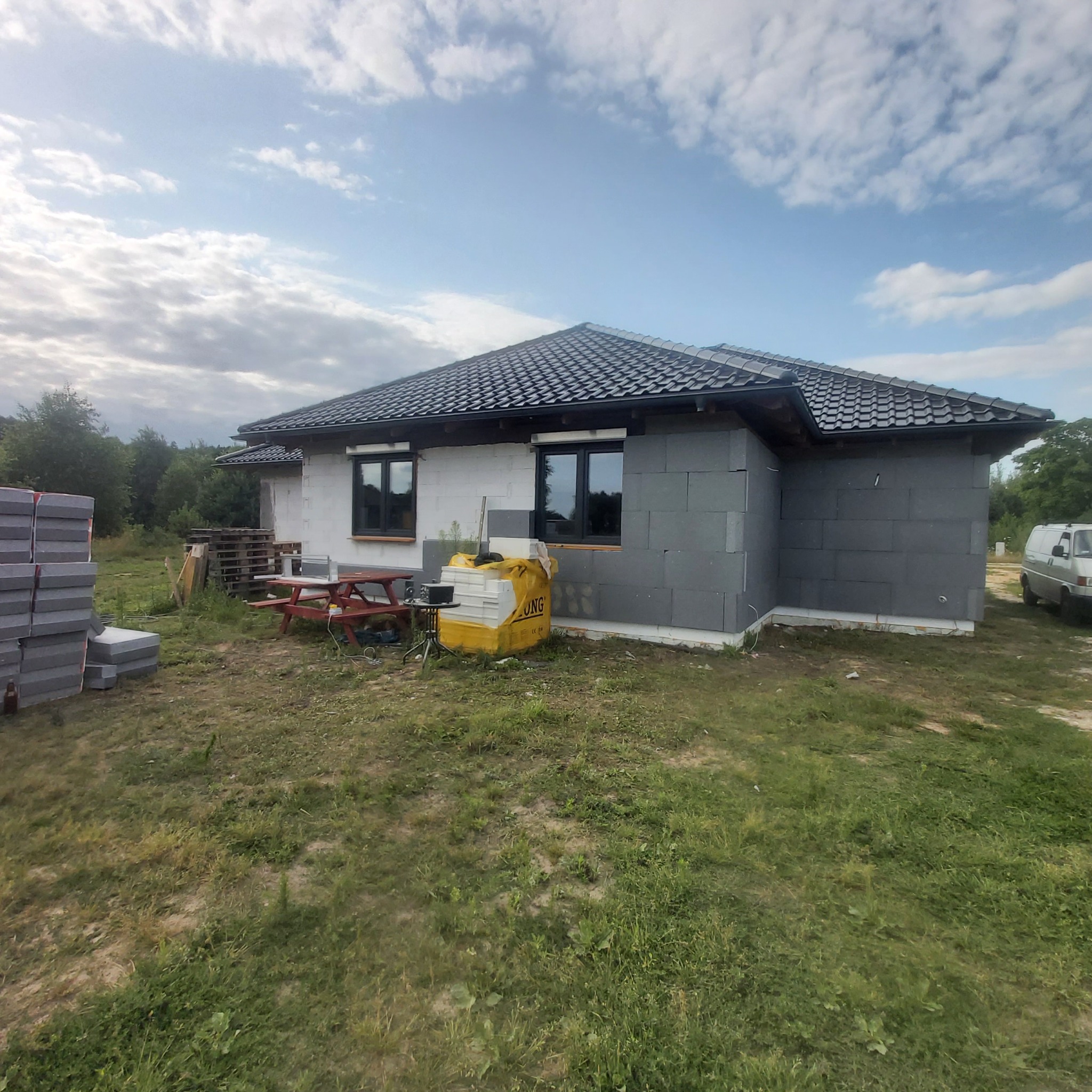 Budynek jednorodzinny w trakcie budowy, widoczne ściany z szarego betonu komórkowego, czarna dachówka, okna z czarnymi ramami, palety z materiałami budowlanymi oraz stół piknikowy na trawiastym...