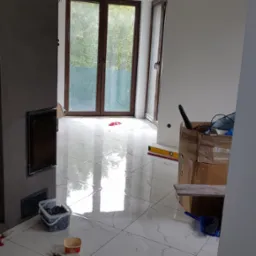 Błyszcząca, biała podłoga z marmurowym wzorem, świeżo ułożona, z widocznymi narzędziami glazurniczymi, poziomnicą i kartonem w tle, w jasnym, przestronnym wnętrzu z dużymi oknami.