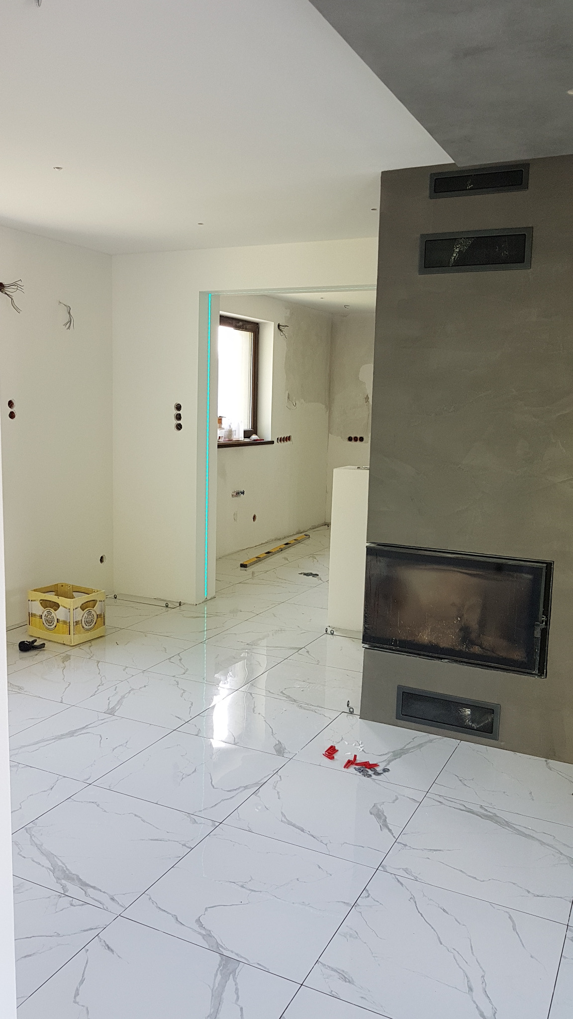 Wnętrze w trakcie prac wykończeniowych: białe marmurowe płytki podłogowe, surowa ściana z kominkiem, widoczne przewody elektryczne i poziomica w przejściu do kolejnego pomieszczenia.