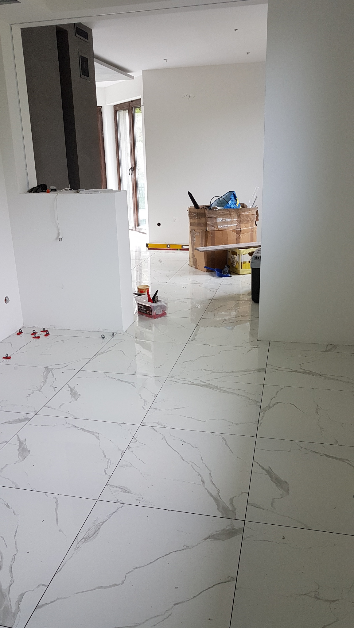 Układanie płytek imitujących marmur na podłodze, widoczne narzędzia glazurnicze, poziomica i kartony w tle, wnętrze w trakcie prac wykończeniowych.