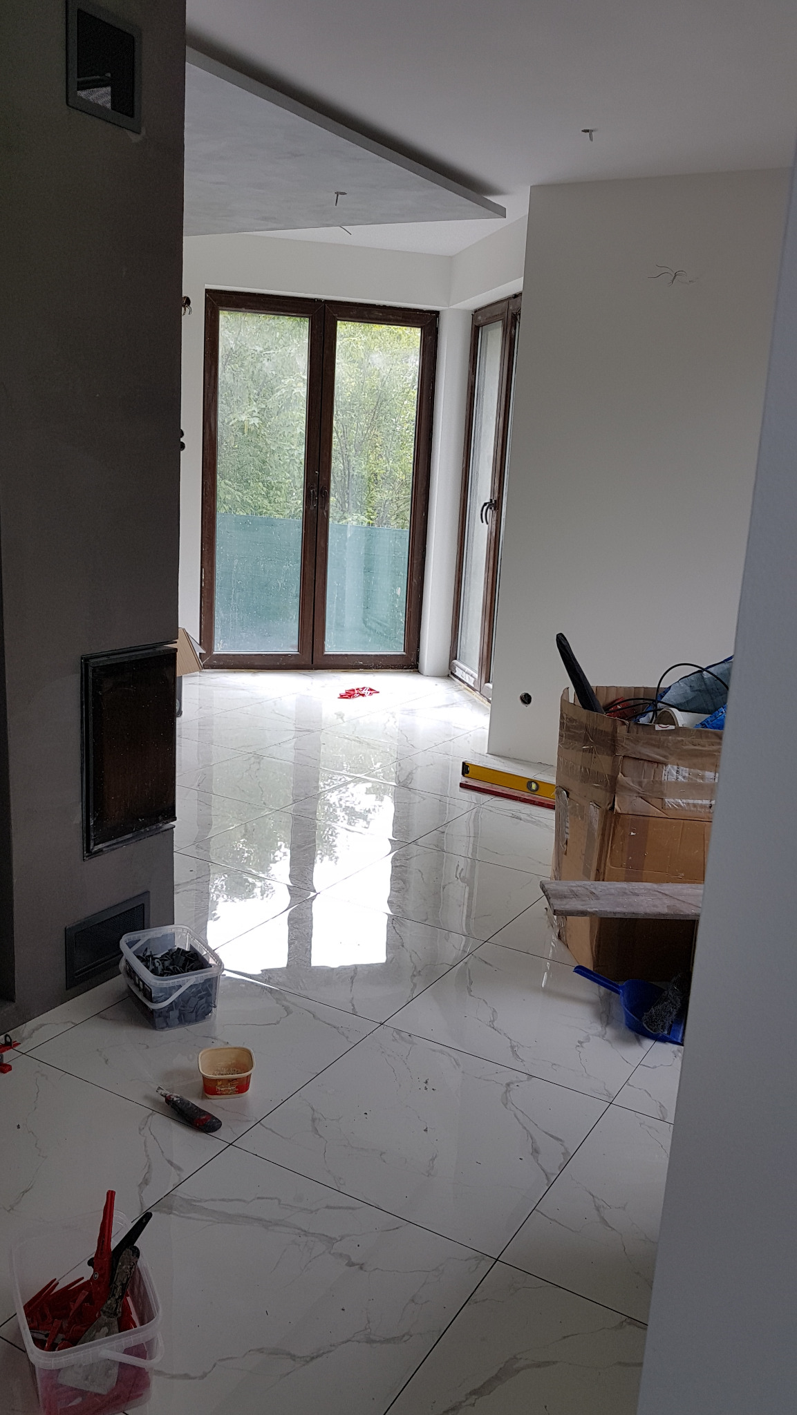 Błyszcząca, biała podłoga z marmurowym wzorem, świeżo ułożona, z widocznymi narzędziami glazurniczymi, poziomnicą i kartonem w tle, w jasnym, przestronnym wnętrzu z dużymi oknami.