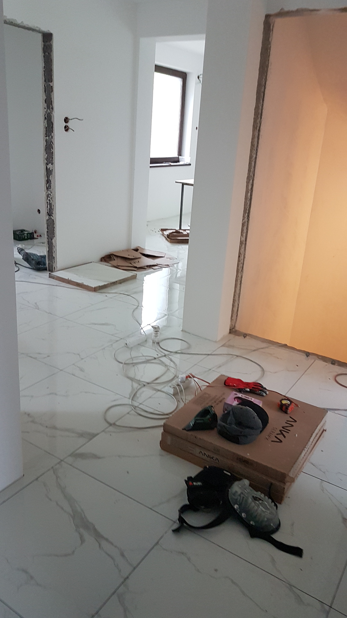 Wnętrze w trakcie remontu: białe marmurowe płytki podłogowe, narzędzia glazurnicze na kartonie, przewody elektryczne rozłożone na podłodze, widoczne otwory drzwiowe w ścianach.
