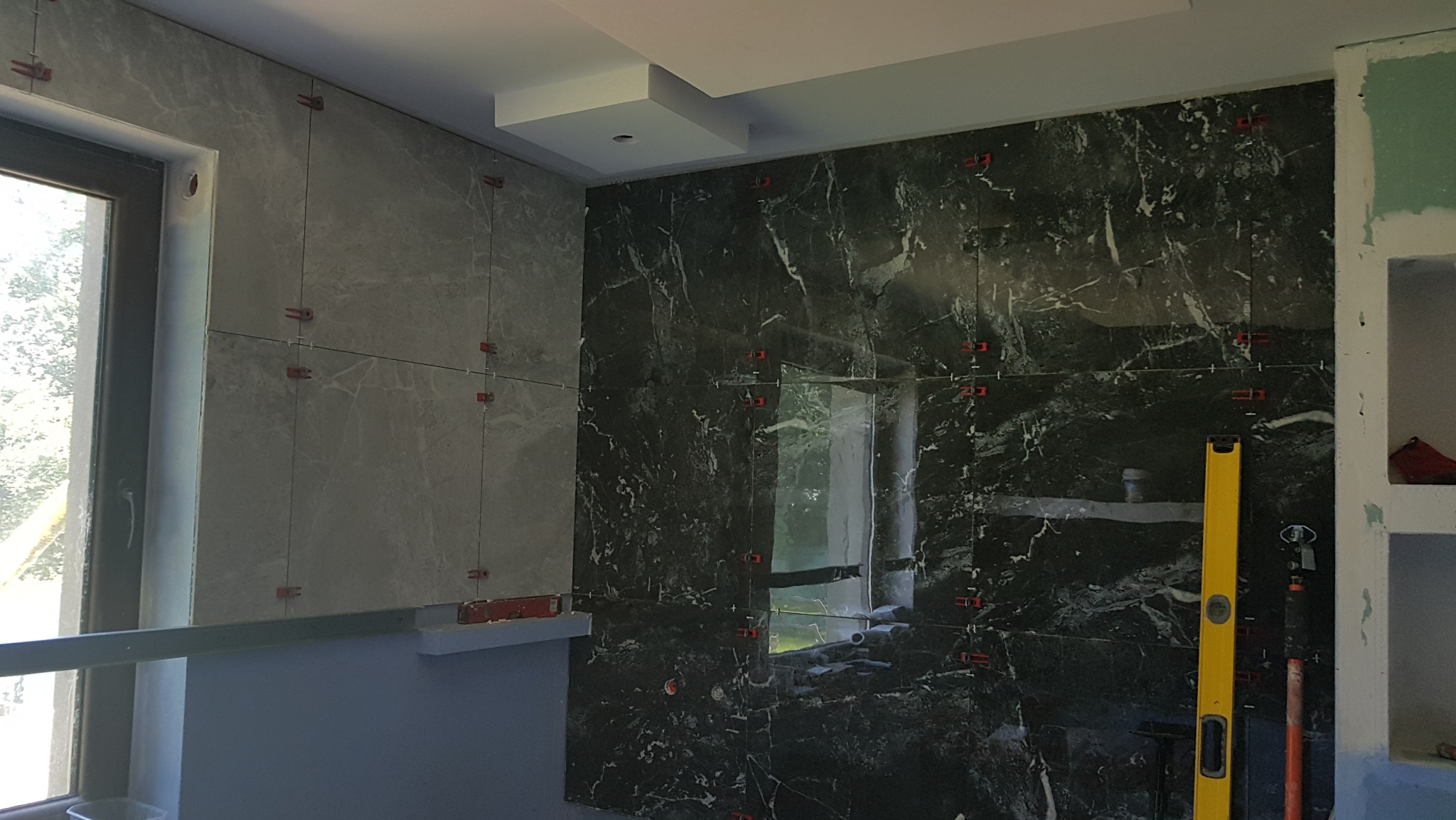 Ściana z częściowo ułożonymi szarymi i czarnymi płytkami imitującymi marmur, z widocznymi krzyżykami dystansowymi i poziomicą, w trakcie prac wykończeniowych.