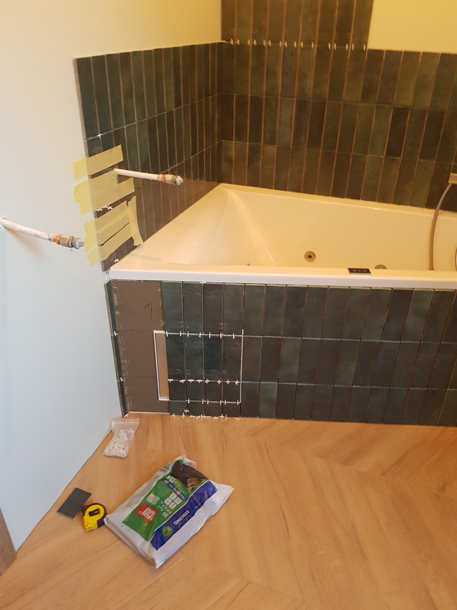 Montaż ciemnozielonych, pionowych płytek nad wanną z hydromasażem, widoczne krzyżyki dystansowe, taśma malarska zabezpieczająca rury, worek z klejem do płytek i miarka na podłodze z paneli.