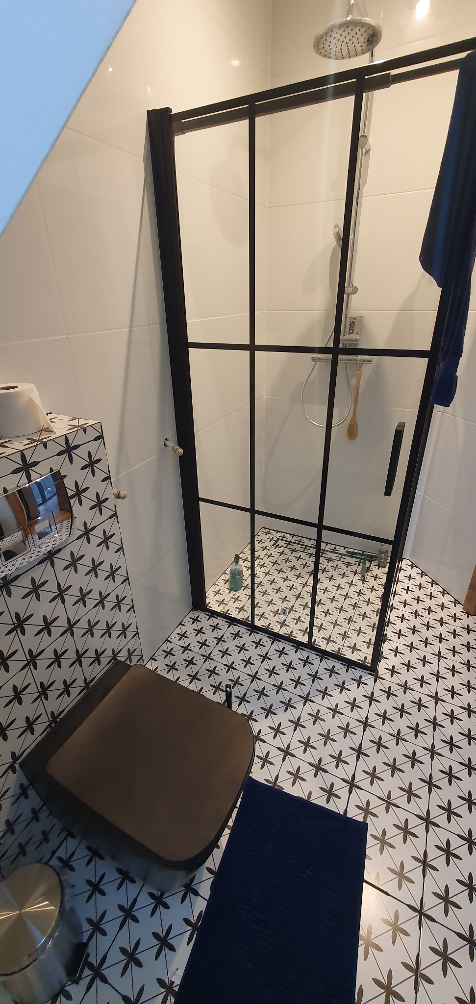 Nowoczesna łazienka z kabiną prysznicową w czarnej ramie, białe płytki z czarnym wzorem, toaleta z deska w kolorze brązowym i niebieski dywanik.