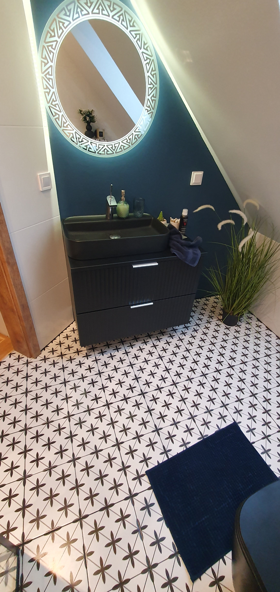 Nowoczesna łazienka na poddaszu: czarna umywalka nablatowa na szafce z ryflowanymi frontami, okrągłe lustro z podświetleniem LED i wzorzyste płytki podłogowe w czarno-białą mozaikę.