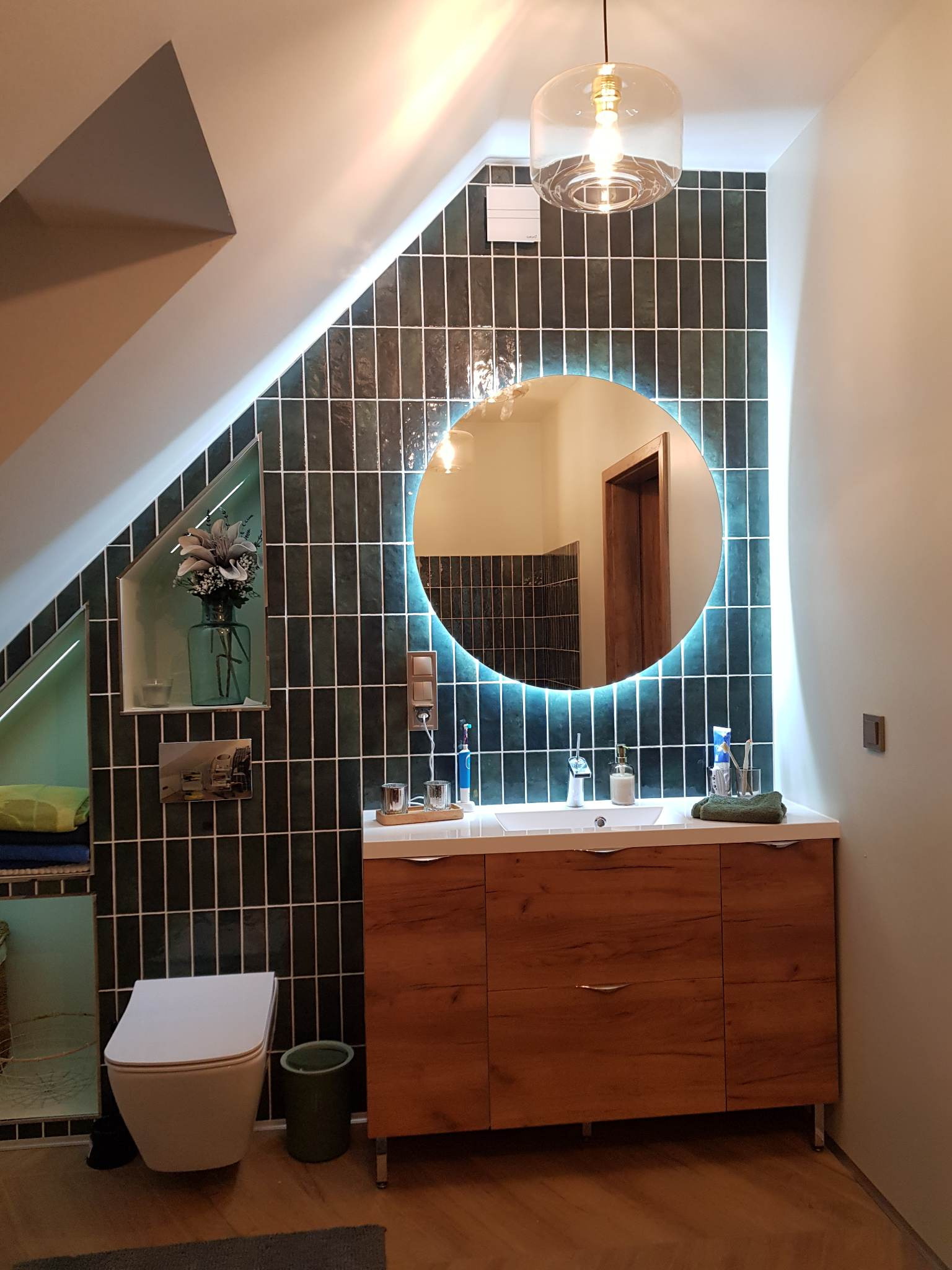 Nowoczesna łazienka na poddaszu z ciemnozielonymi, pionowo ułożonymi płytkami, okrągłym lustrem z podświetleniem LED, drewnianą szafką pod umywalką i wiszącą lampą z przezroczystym kloszem.