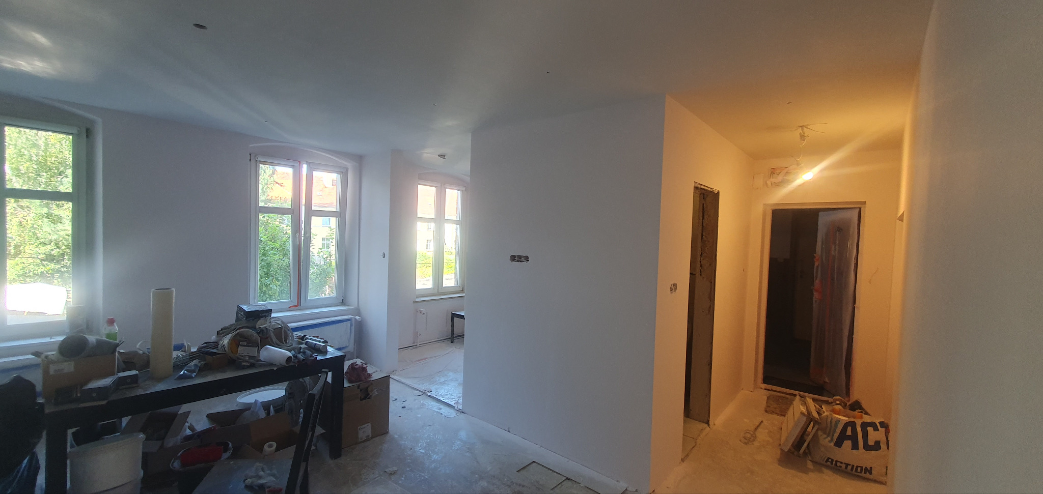 Wnętrze w trakcie prac wykończeniowych: świeżo pomalowane ściany na biało, widoczne okna, przygotowane narzędzia i materiały budowlane na stole, surowy stan podłogi.