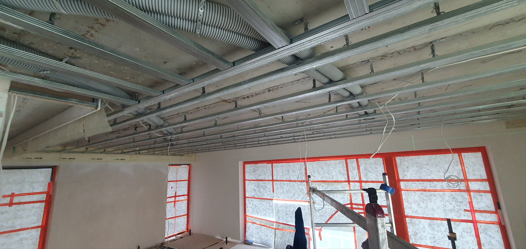Sufit podwieszany z metalową konstrukcją i rurami wentylacyjnymi w surowym stanie, okna zabezpieczone folią ochronną, drabina w pomieszczeniu.