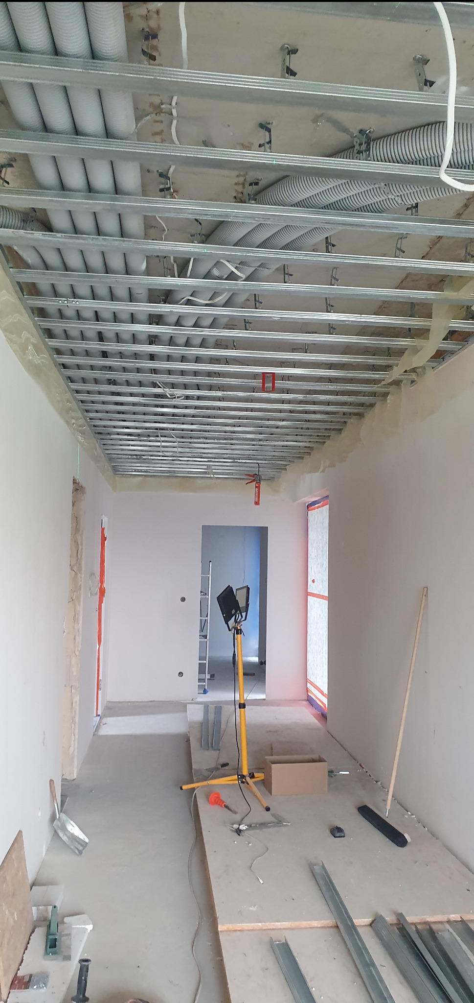 Sufit podwieszany w trakcie montażu: metalowa konstrukcja, rury wentylacyjne. Widoczne narzędzia i oświetlenie budowlane. Wnętrze w stanie surowym.