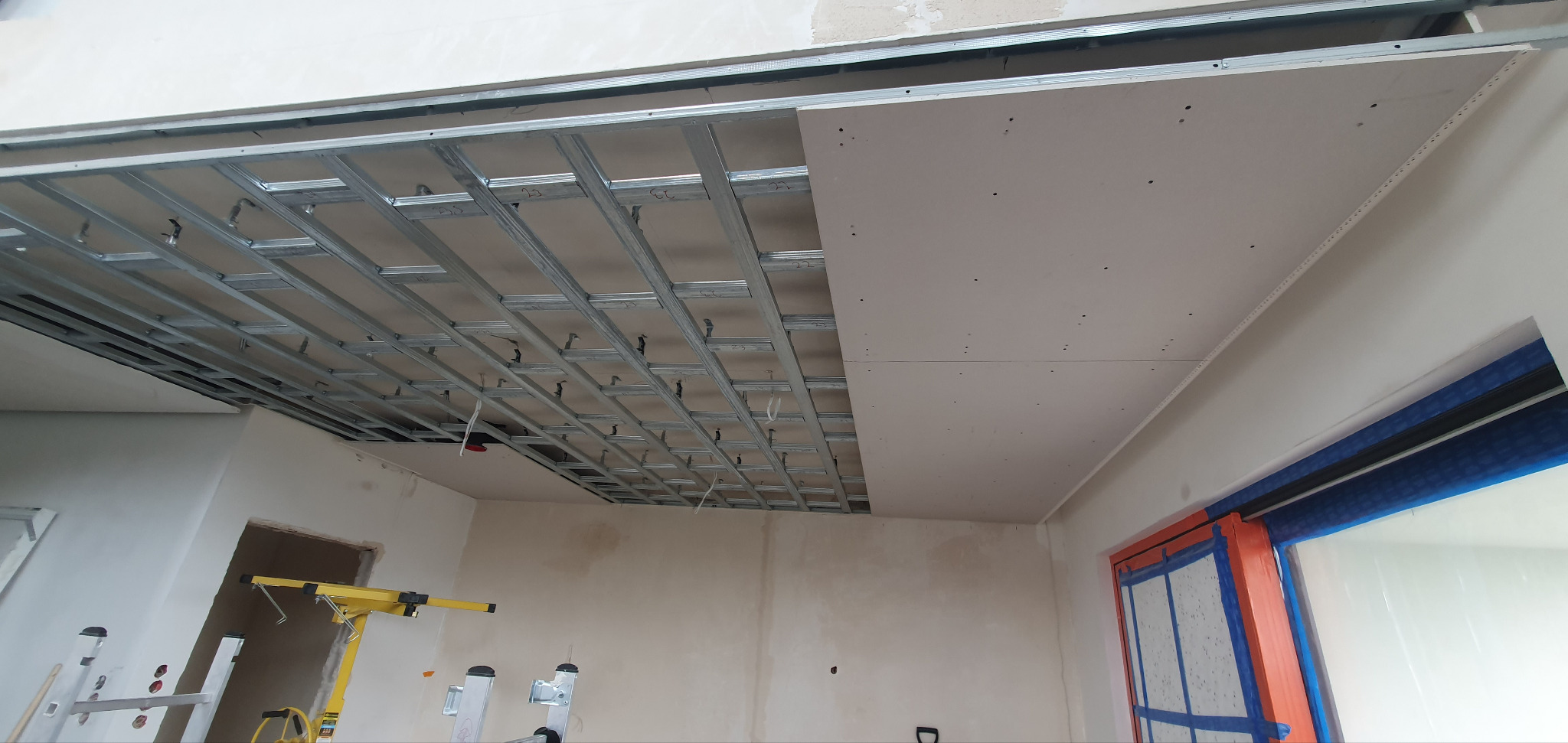 Montaż sufitu podwieszanego: metalowa konstrukcja i płyty gipsowo-kartonowe w trakcie instalacji. Widoczne narzędzia i elementy wykończeniowe. Prace wykończeniowe w pomieszczeniu.