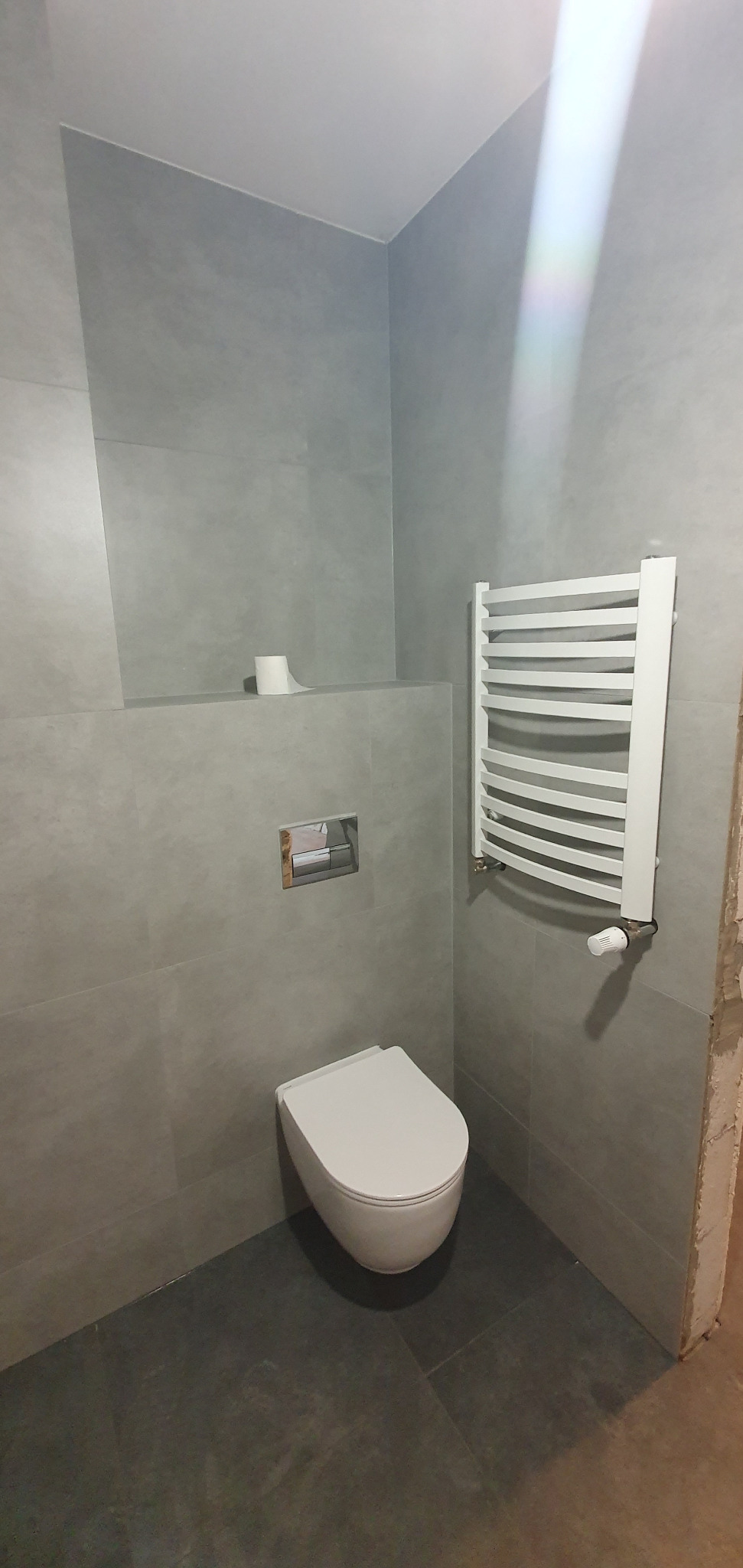 Nowoczesna łazienka z szarymi płytkami, podwieszaną toaletą i białym grzejnikiem drabinkowym. Minimalistyczny design, widoczne detale wykończenia.