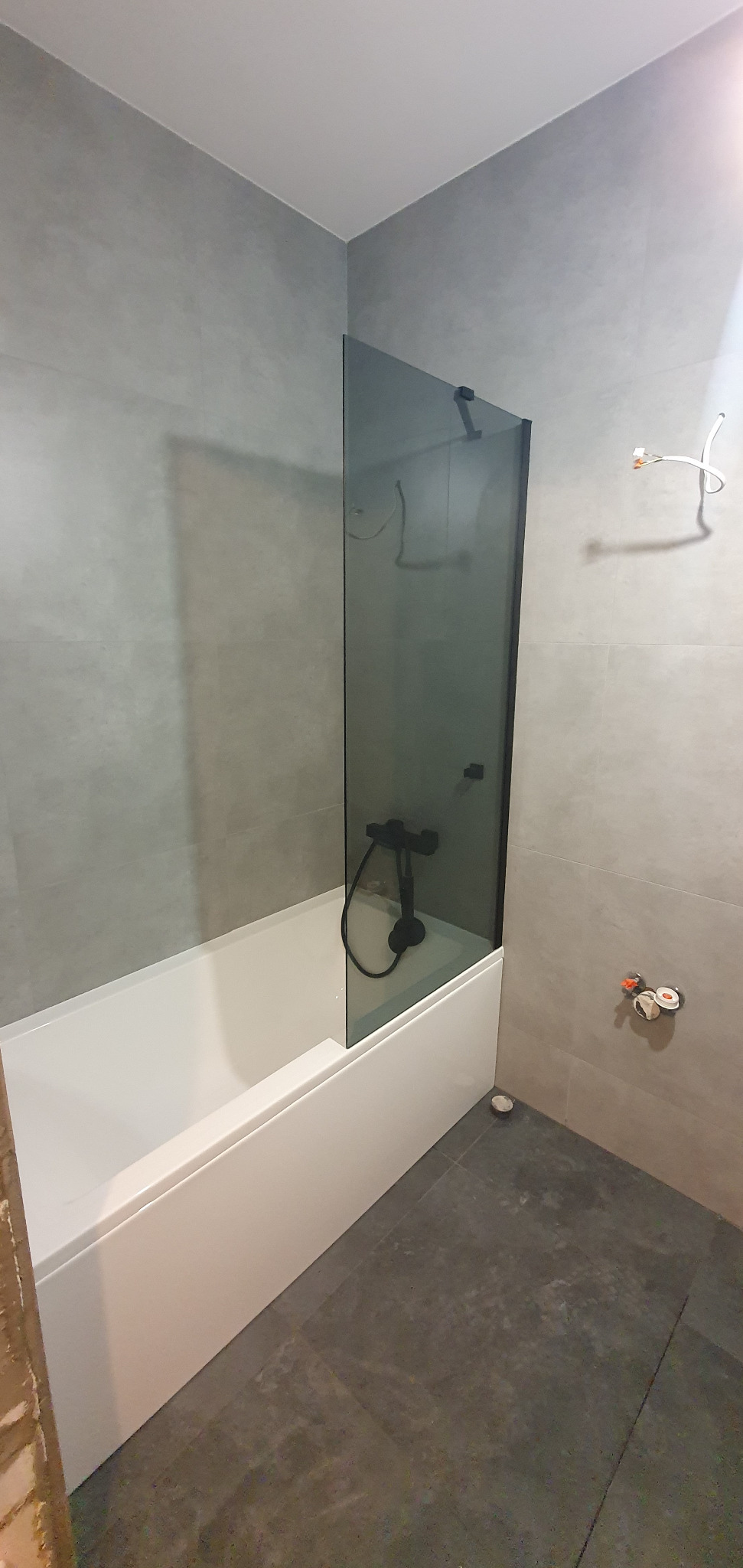 Nowoczesna łazienka z szarymi płytkami, białą wanną i czarną armaturą. Kabina prysznicowa z ciemnego szkła. Minimalistyczny design.