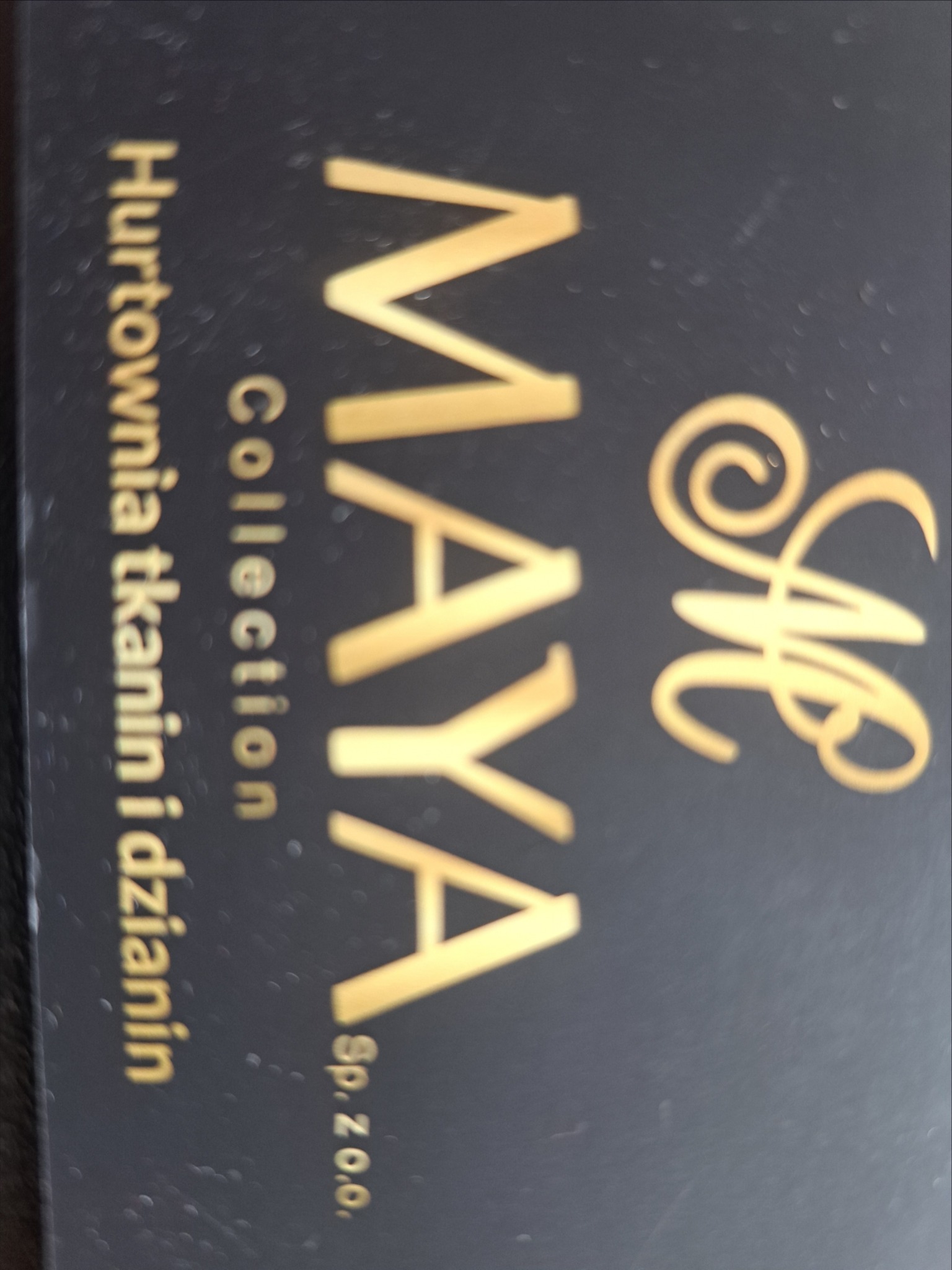Wizytówka firmy z nazwą Maya Collection, specjalizującej się w tkaninach i dzianinach, złote napisy na ciemnym tle.