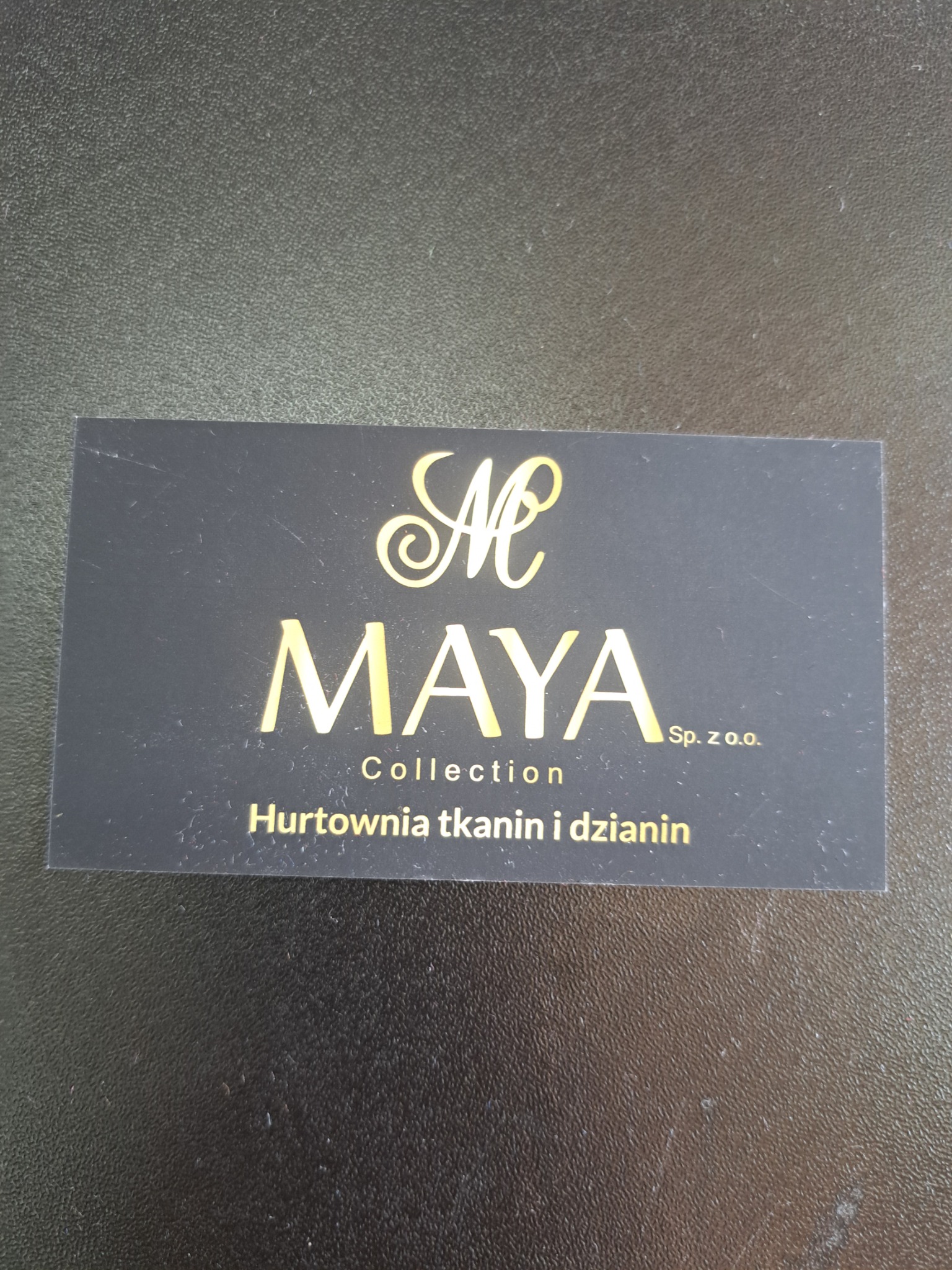 Wizytówka firmy Maya Collection z Rzgowa, specjalizującej się w tkaninach i dzianinach. Czarny kartonik ze złotym napisem na tle ciemnej powierzchni.