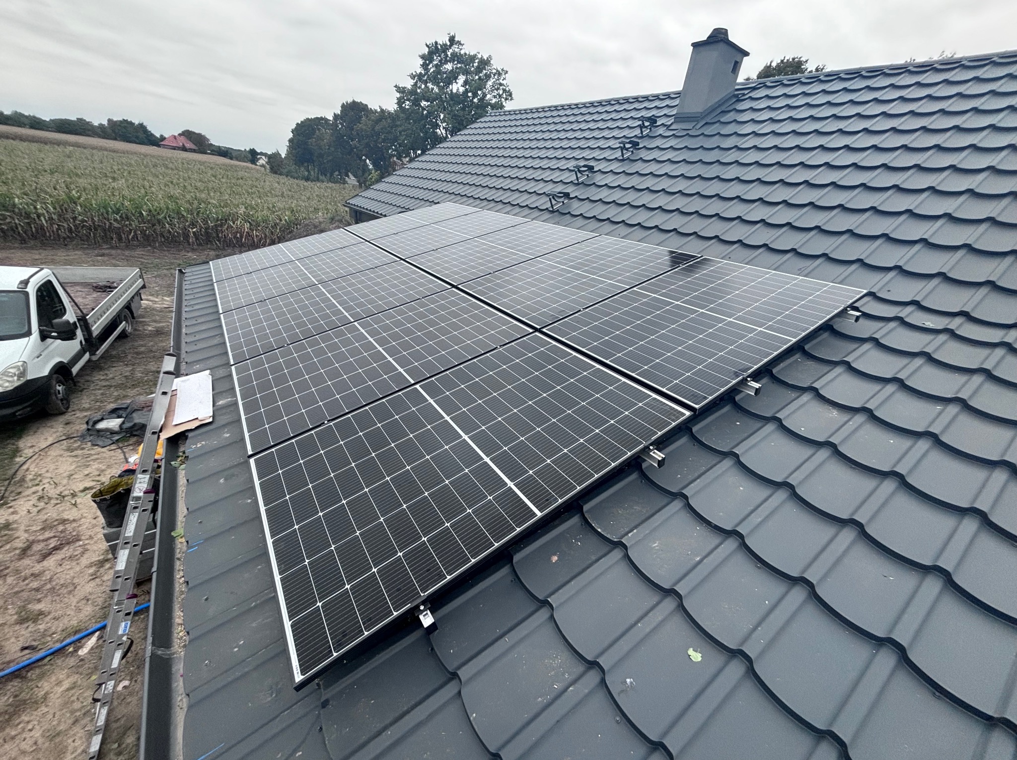 Panel słoneczny na grafitowym dachu z blachodachówki, widok z góry, drabina oparta o rynnę, w tle pole kukurydzy i zabudowania.