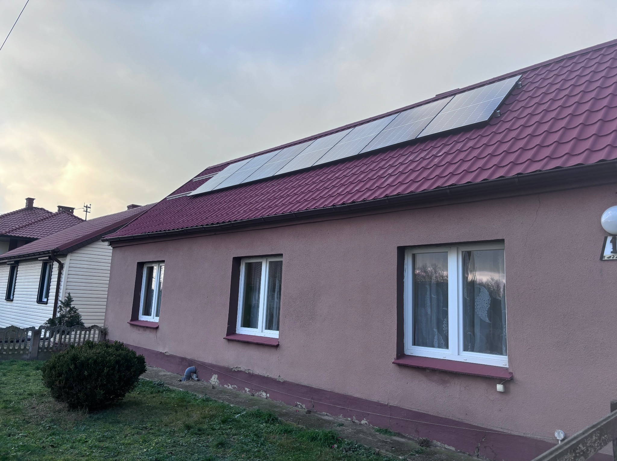 Dom jednorodzinny z panelami słonecznymi na bordowym dachu z dachówki ceramicznej, widok z niskiej perspektywy w pochmurny dzień.